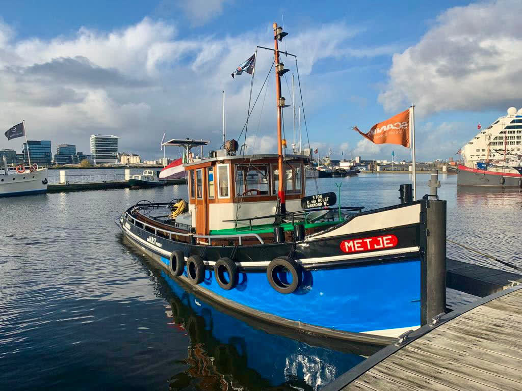 Sleepboot Metje afgemeerd in een haven aan het IJ, met de skyline van Amsterdam op de achtergrond