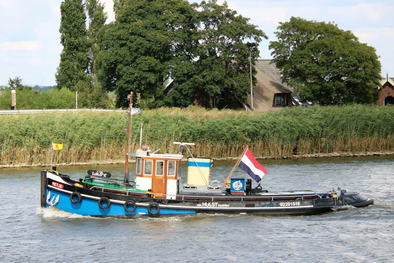 Sleepboot Metje vaart langs een rietoever bij Warmond met de Nederlandse vlag in top