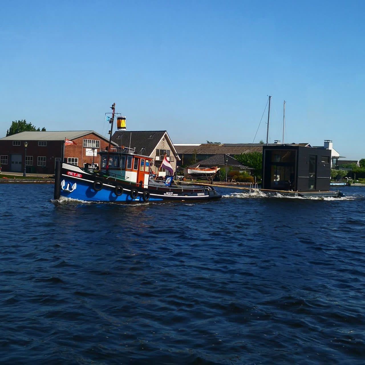 Sleepboot Metje versleept een moderne woonark langs een jachtwerf bij Warmond