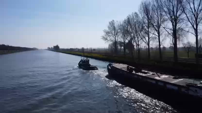 Sleepboot Metje sleept een volgeladen vrachtschip over een kanaal, gezien vanuit de lucht